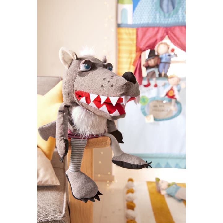 Eine große graue Wolfspuppe mit rotem Mund und weißen Zähnen sitzt auf dem Arm eines beigen Sofas. Im Hintergrund befindet sich ein buntes Stoffpuppentheater mit menschlichen Puppen und einem dekorativen Teppich auf dem Boden.