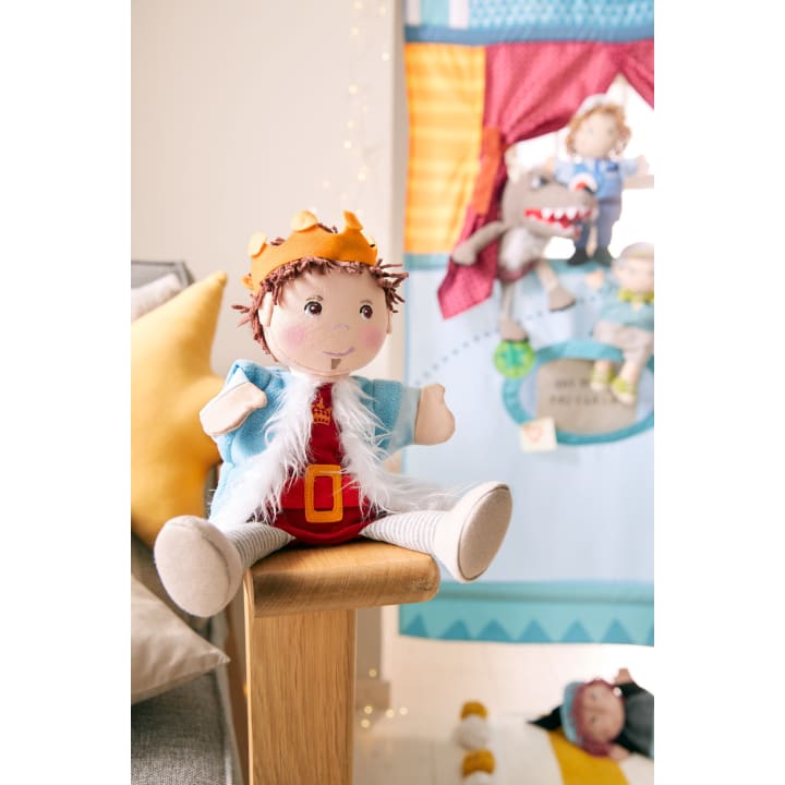 Eine Plüschpuppe, gekleidet als König mit Krone, blauem Umhang und rotem Outfit, sitzt auf einer Holzfläche. Im Hintergrund befinden sich weitere Puppen und eine bunte Stoffszene, die an der Wand hängt.