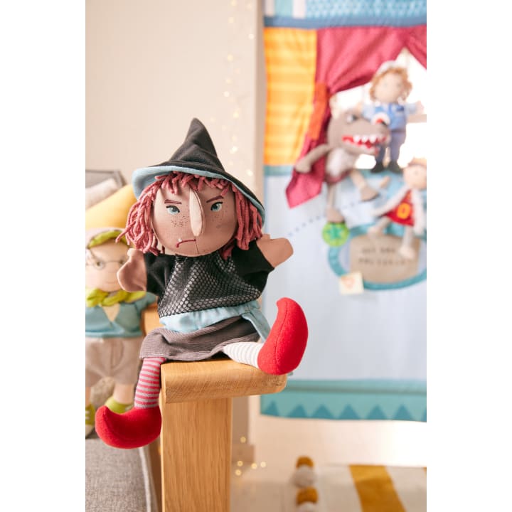 Eine Plüschhexenpuppe mit rotem Garnhaar, einem schwarzen Hut, roten Schuhen und einer gestreiften Socke sitzt auf dem Rand einer Holzfläche. Im unscharfen Hintergrund sind weitere Plüschpuppen und ein bunter Stoffhintergrund zu sehen.