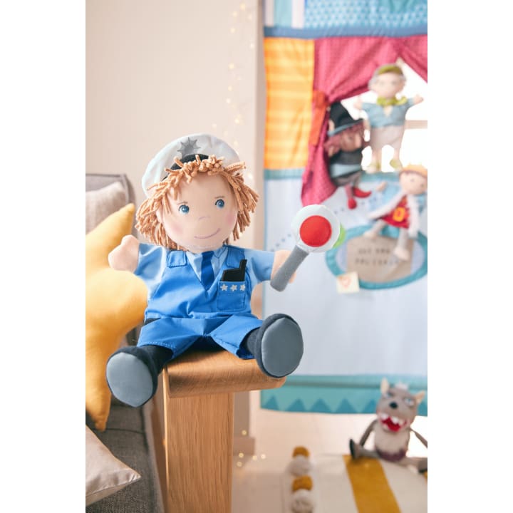 Eine als Polizist verkleidete Plüschpuppe sitzt auf der Armlehne eines Holzstuhls und hält einen Spielzeugschlagstock. Im Hintergrund ist ein buntes Puppentheater mit anderen Plüschpuppen und Spielzeug zu sehen.