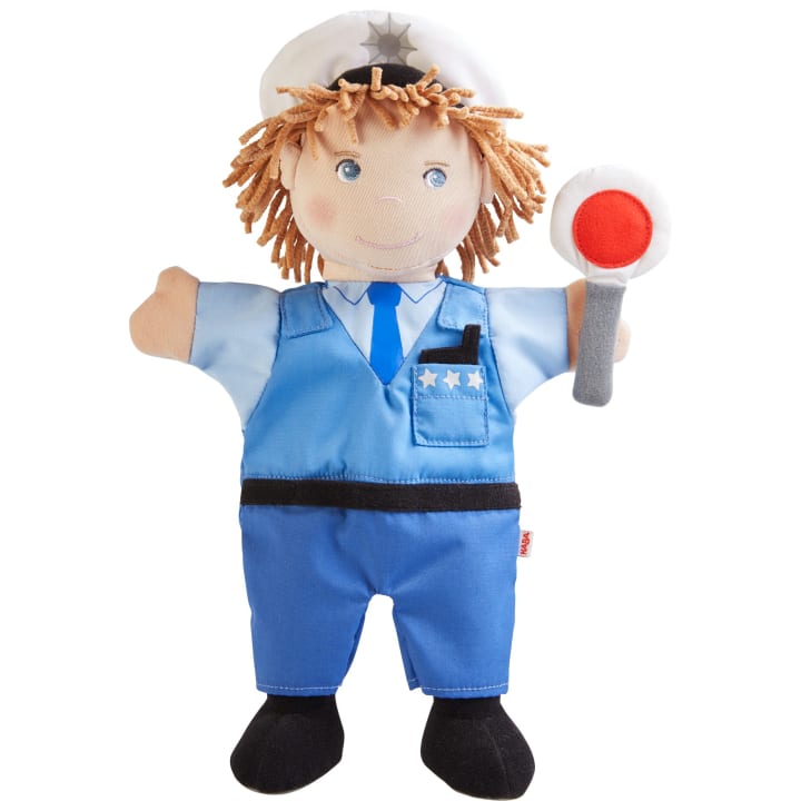 Eine weiche Stoffpuppe in einer blauen Polizeiuniform mit schwarzen Schuhen, einer weißen Polizeimütze und drei Sternen auf der Hemdtasche, die einen grauen Stock mit einem rot-weißen Kreis am Ende hält.