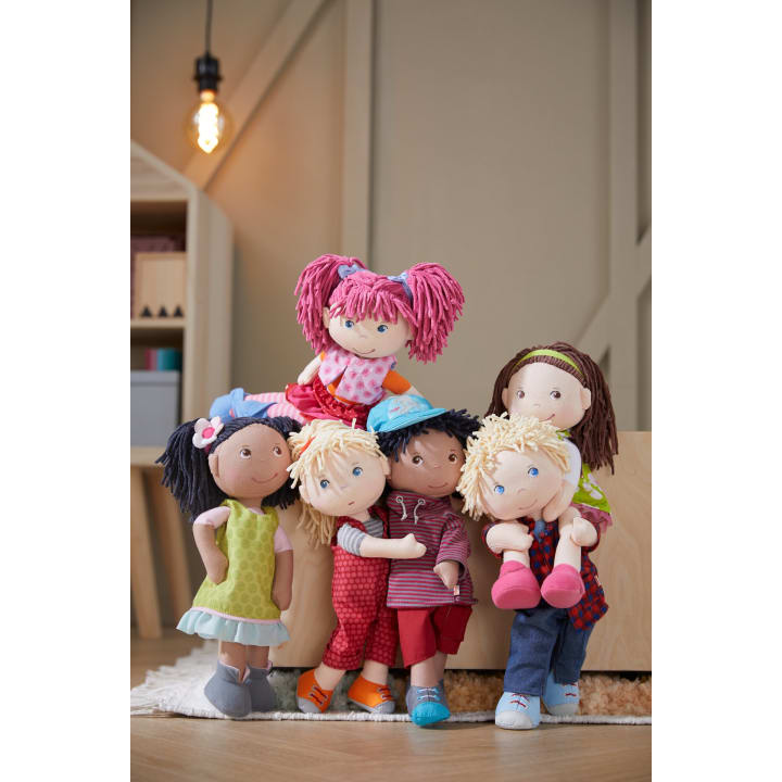 Sechs Stoffpuppen mit farbenfroher Kleidung und unterschiedlichen Haarfarben und Frisuren sind in einem Raum vor Holzmöbeln und einem Bücherregal unter einer beleuchteten Hängelampe gruppiert. Die Puppen sind in freundlichen Posen angeordnet.