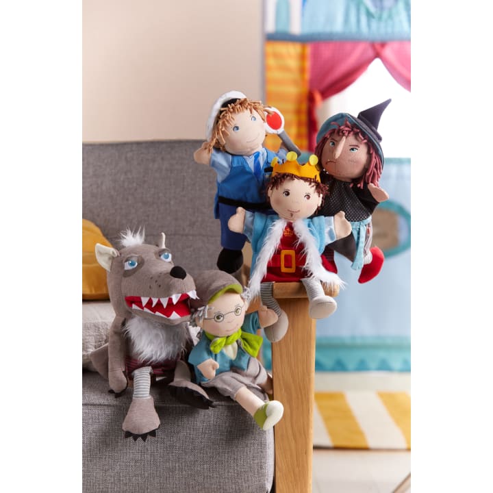 Fünf Plüschpuppen, darunter ein Wolf, eine ältere Person mit Brille, ein Kind mit Krone, eine Figur mit schwarzem Hut und eine Person in Blau, sind auf einer Couch und einer Armlehne in einem farbenfrohen Raum angeordnet.