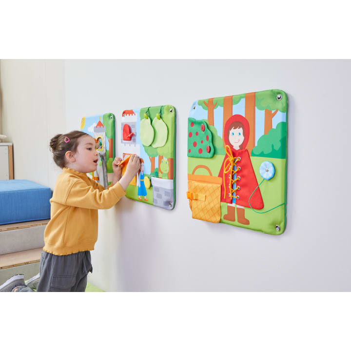 Ein junges Mädchen interagiert mit bunten, strukturierten Aktivitätstafeln, die an einer Wand angebracht sind, darunter eine mit einem Rotkäppchen-Motiv und eine andere mit hängenden Filzfrüchten.