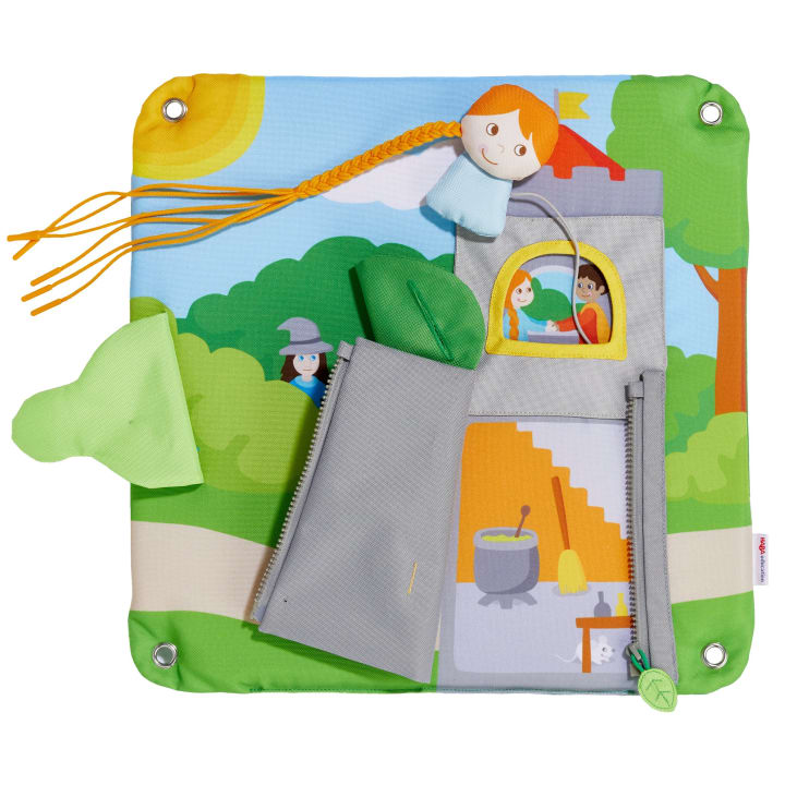 Ein farbenfrohes Activity Board aus Stoff mit einer Burgszene, die eine Tür mit Reißverschluss, eine kleine Hexenfigur, einen weichen Besen und ein Mädchen mit orangefarbenem Garnhaar zeigt. Zwei Kinder und ein Besen sind in einem Fenster abgebildet.