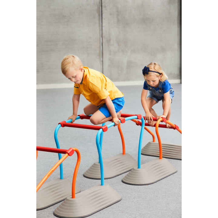 Zwei kleine Kinder klettern über Plastikhürden mit blauen und roten Stäben in einem Innenspielbereich mit grauem Boden und Betonwand.