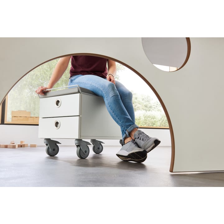 Eine Person in Jeans und grauen Turnschuhen sitzt auf einem weißen Rollschrank mit zwei Schubladen. Das Foto ist durch einen runden Ausschnitt in einer weißen Struktur aufgenommen. Im Hintergrund sind Holzblöcke und große Fenster zu sehen.