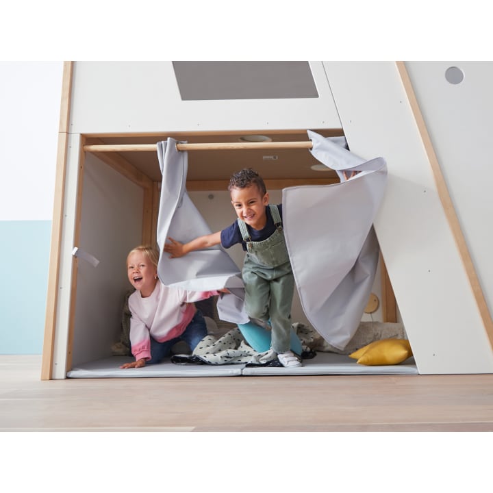 Zwei kleine Kinder, ein Mädchen und ein Junge, krabbeln und lachen, als sie aus einem Spielzelt mit weißen Vorhängen in einem hell erleuchteten Raum herauskommen. Im Inneren des Zeltes sind ein Kissen und eine Decke zu sehen.