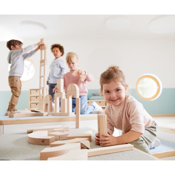 Vier kleine Kinder spielen mit Holzbausteinen in einem hellen, modernen Raum. Ein Mädchen lächelt in die Kamera, während es Klötze in der Hand hält, während die anderen sich darauf konzentrieren, Klötze auf Tischen zu stapeln und anzuordnen.