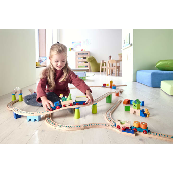 Ein junges Mädchen sitzt auf einem hellen Holzboden und spielt mit einer Holzeisenbahn mit Schienen, Brücken und kleinen Gebäuden in einem hellen, aufgeräumten Zimmer mit kindgerechten Möbeln und bunten Kissen.