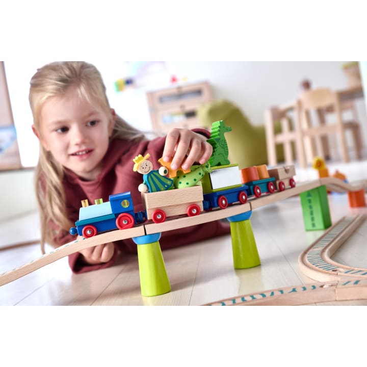 Ein junges Mädchen liegt auf dem Boden und stellt eine hölzerne Spielzeugfigur auf eine hölzerne Spielzeugeisenbahn, die durch bunte Blöcke erhöht ist. Der Zug hat mehrere Waggons mit verschiedenen Spielzeugartikeln im Inneren.
