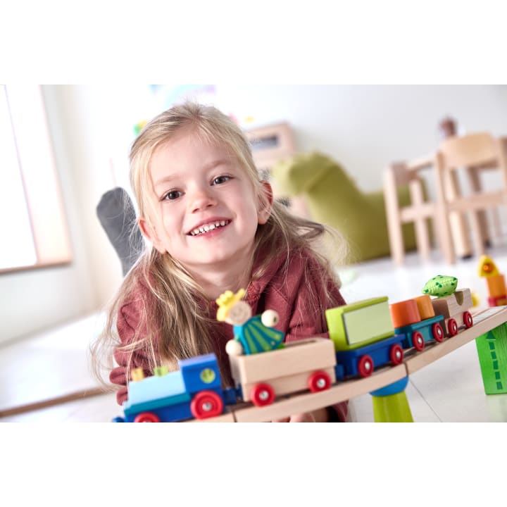 Ein kleines Kind mit langen blonden Haaren lächelt, während es mit einem bunten hölzernen Spielzeugeisenbahn auf einer Strecke drinnen spielt. Der Hintergrund zeigt ein Spielzimmer mit Möbeln und weichem Tageslicht.