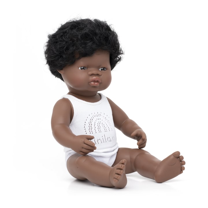 Eine dunkelhäutige Babypuppe mit lockigem schwarzem Haar sitzt aufrecht und trägt einen weißen ärmellosen Strampler mit einem kleinen Muster auf der Vorderseite vor einem schlichten weißen Hintergrund.