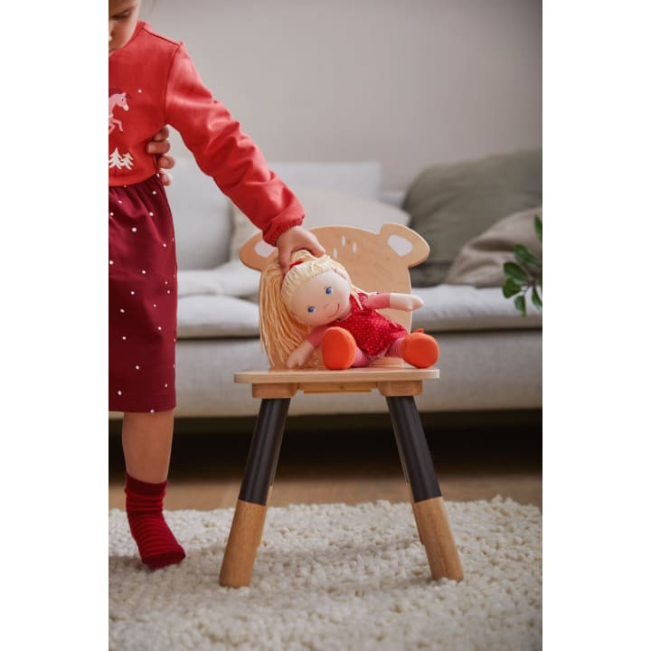 Ein Kind in einem roten Kleid und roten Socken setzt eine blonde Puppe in einem roten Outfit auf einen kleinen Holzstuhl mit tierförmigen Details. Die Szene spielt in einem Wohnzimmer mit einem weißen Teppich und einem Sofa im Hintergrund.