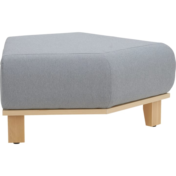 Ein moderner, hellgrauer, gepolsterter Hocker mit niedrigem, kantigem Design. Er hat einen Holzsockel und drei Holzbeine, wobei der Hocker auf einem weißen Hintergrund steht.