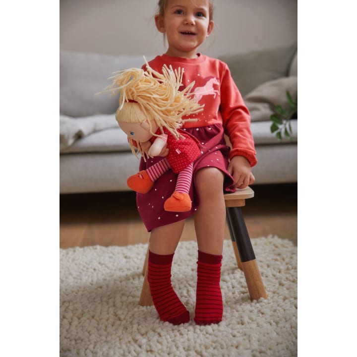 Ein kleines Kind in einem rosa-roten Kleid und roten Socken sitzt auf einem kleinen Holzschemel und hält eine Puppe mit blondem Garnhaar und rotem Outfit. Im Hintergrund sind eine beigefarbene Couch und ein Teppich zu sehen.