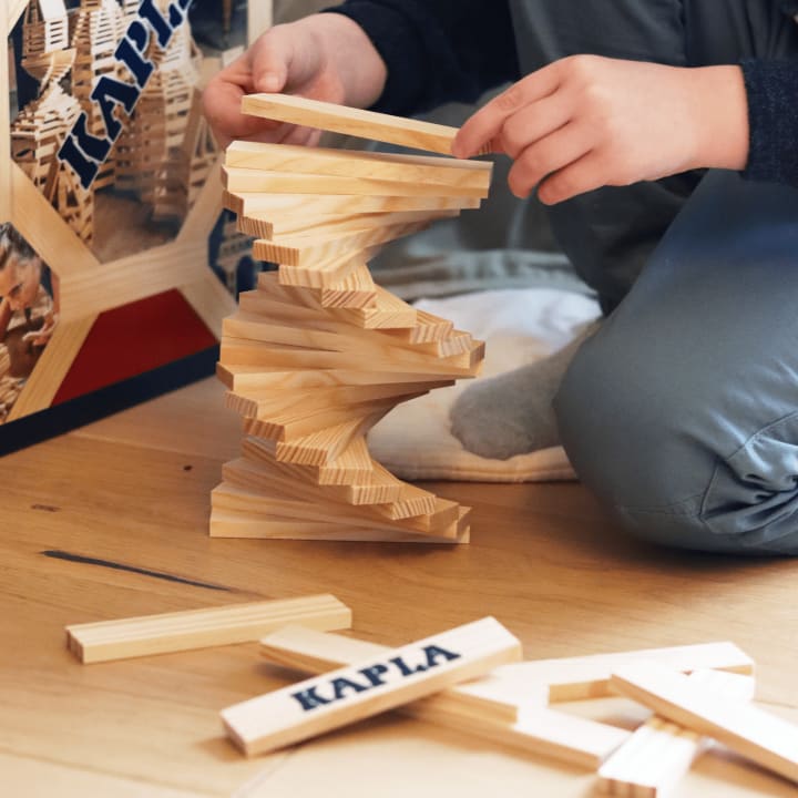 Kinderhände bauen einen spiralförmigen Turm aus Kapla-Holzbrettern auf einem Holzboden, mit weiteren Brettern und einer Kapla-Box im Hintergrund.