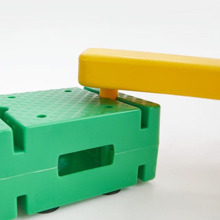 Nahaufnahme eines gelben Kunststoffbalkens, der mit einem grünen Kunststoffsockel mit strukturierter Oberfläche und rechteckigem Loch verbunden ist; beide Teile sind wahrscheinlich Teil eines modularen Spielzeugs oder Bausatzes.