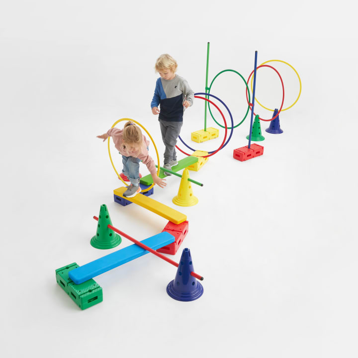 Zwei kleine Kinder balancieren und laufen über einen bunten Hindernisparcours aus Plastikbalken, Kegeln, Reifen und Blöcken auf weißem Hintergrund. Der Parcours besteht aus verschiedenen Formen und leuchtenden Grundfarben.