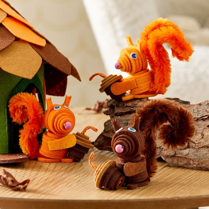 Drei Eichhörnchenfiguren aus Filz und Pfeifenreinigern sind auf einer Holzfläche neben einem Baumhaus aus Filz und einem Stück Baumrinde aufgestellt. Die Eichhörnchen sind orange und braun und halten jeweils eine Eichel aus Filz.
