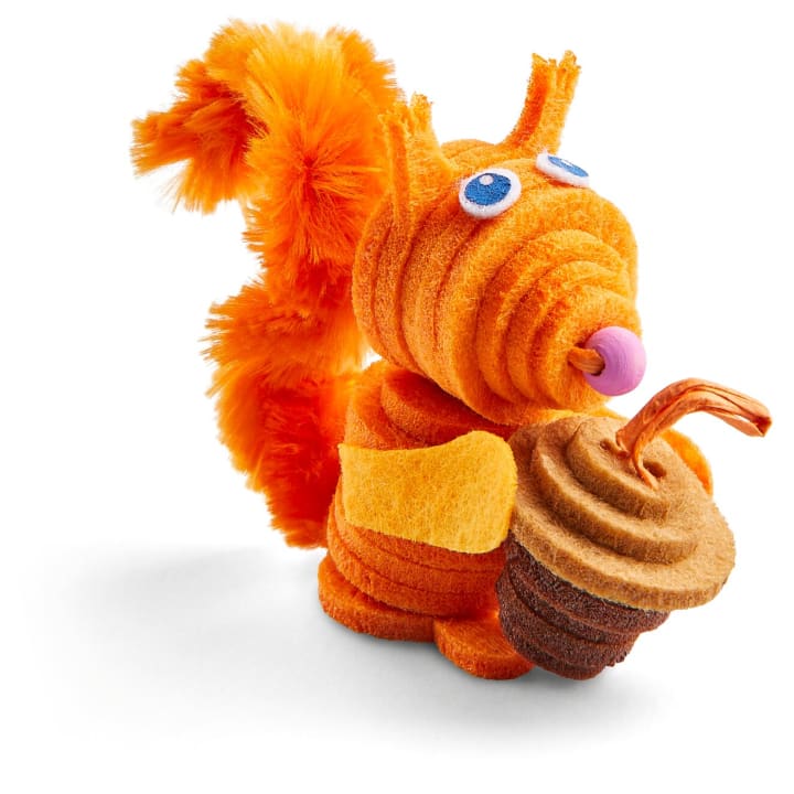 Eine kleine Eichhörnchenskulptur aus orangefarbenen und braunen Chenille-Pfeifenputzern mit blauen und weißen Filzaugen, die eine braune Eichel halten, auf weißem Hintergrund.