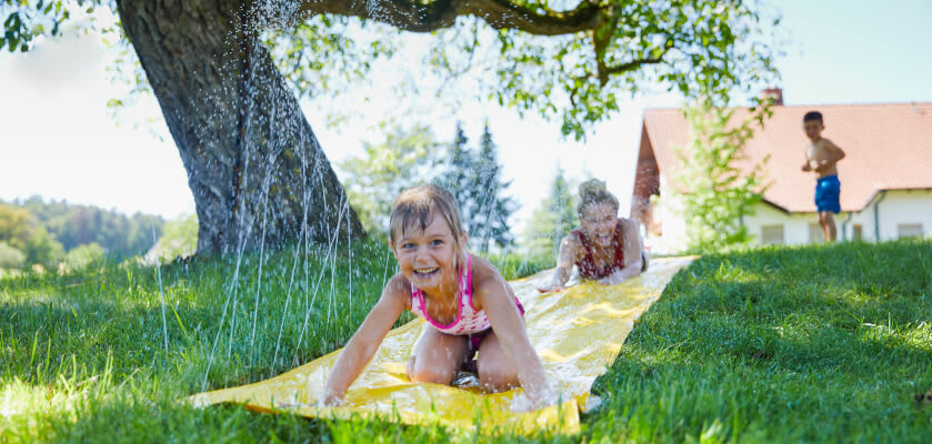 Wasserspiele mit Kindern: Glückliche Kinder auf einer Wasserrutsche im Garten