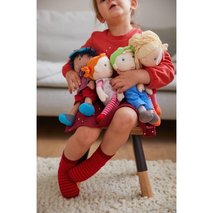 Ein kleines Kind in einem roten Kleid und rot gestreiften Socken sitzt auf einem kleinen Hocker und hält vier bunte Stoffpuppen auf dem Schoß. Im Hintergrund sind ein graues Sofa und ein cremefarbener Teppich zu sehen.