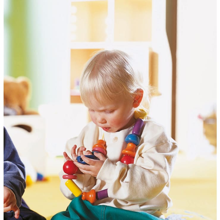 Ein kleines Kind mit hellem Haar, das ein cremefarbenes Hemd und eine Kette aus großen, bunten Perlen trägt, sitzt im Haus und konzentriert sich auf das Stapeln oder Untersuchen ähnlicher Spielzeugteile in seinen Händen.