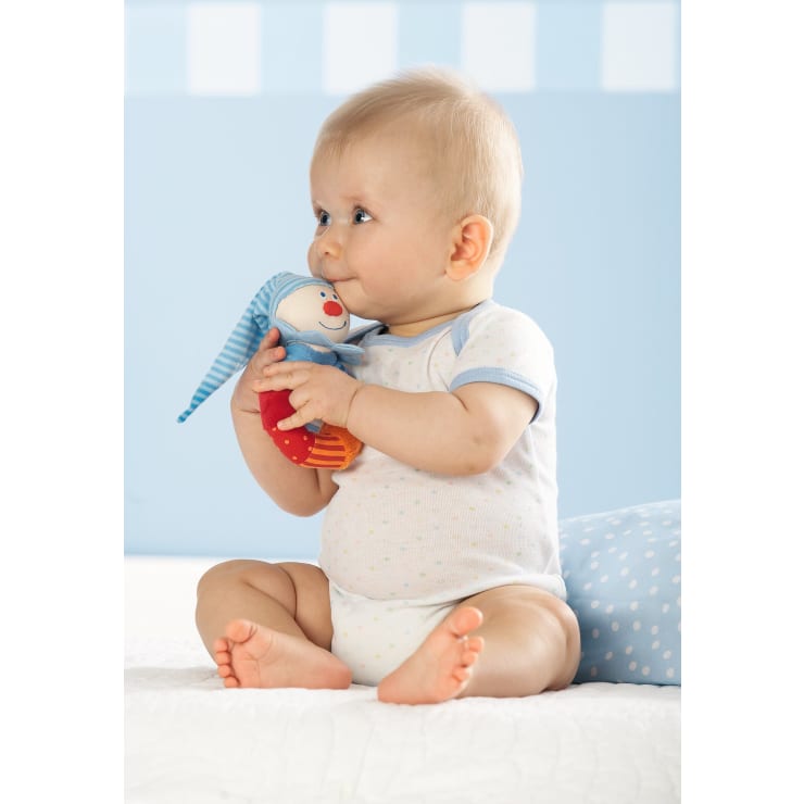 Ein Baby in einem weißen Body mit hellblauem Rand sitzt auf einem Bett und hält ein Stofftier mit einer blauen Mütze. Das Baby blickt zur Seite, und der Hintergrund ist hellblau mit einem gepunkteten Kissen.