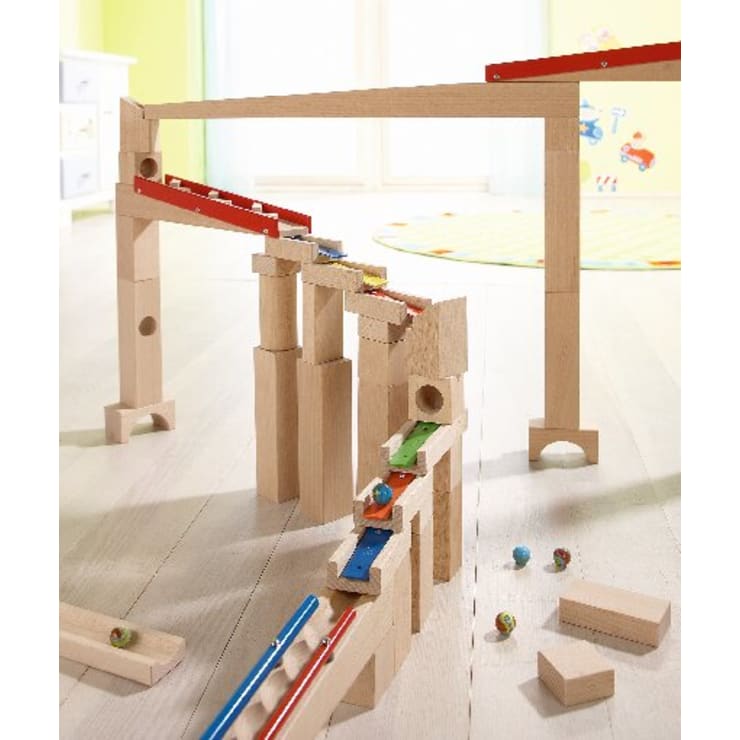 Eine hölzerne Murmelbahn ist auf einem hellen Boden aufgebaut, wobei Murmeln auf der Bahn und in der Nähe sichtbar sind. Im Hintergrund befinden sich ein Teppich, ein Fenster und eine teilweise sichtbare Kinder-Wandmalerei.