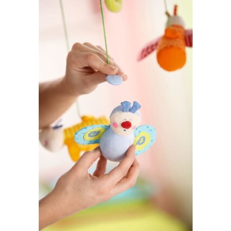 Die Hände einer Person halten ein blaues vogelförmiges Plüschtier, das an einer grünen Schnur als Teil eines Babymobiles hängt. Andere bunte Plüschtiere sind im Hintergrund zu sehen.