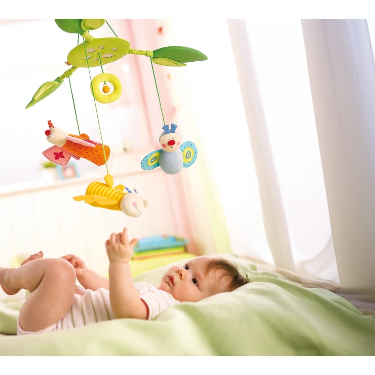 Ein auf einem Bett liegendes Baby streckt seine Hand nach einem bunten Mobile mit Plüschtierfiguren aus, das in einem hell erleuchteten Raum über ihm hängt.