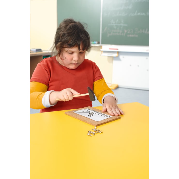 Ein Kind mit dunklen Haaren und einem rot-orangefarbenen Hemd benutzt einen kleinen Hammer, um Stecknadeln in eine Korkplatte auf einem gelben Tisch zu stecken. Im Hintergrund sind eine Kreidetafel und die Möbel des Klassenzimmers zu sehen.