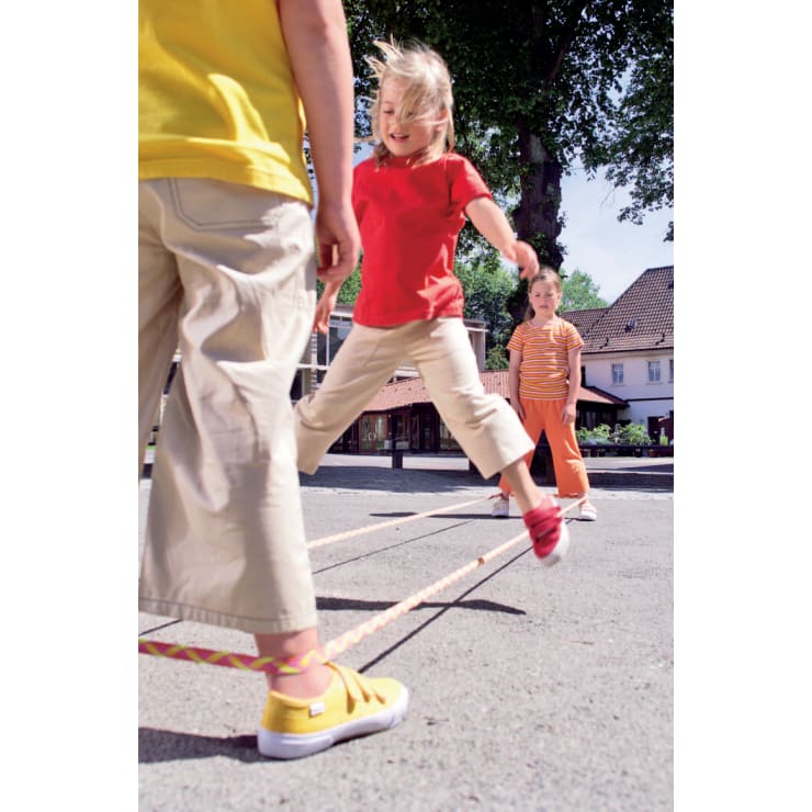 Drei Kinder spielen im Freien ein Springspiel mit einem langen Gummiband. Ein Kind springt gerade über das Band, ein anderes steht innerhalb des Gummibandes und ein drittes Kind schaut im Hintergrund zu.