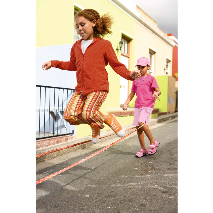 Zwei Kinder spielen draußen auf einer Straße Seilspringen. Ein Kind, das eine orangefarbene Jacke und eine gestreifte Hose trägt, springt, während das andere, das in Rosa gekleidet ist, das Seil dreht. Im Hintergrund sind bunte Gebäude zu sehen.