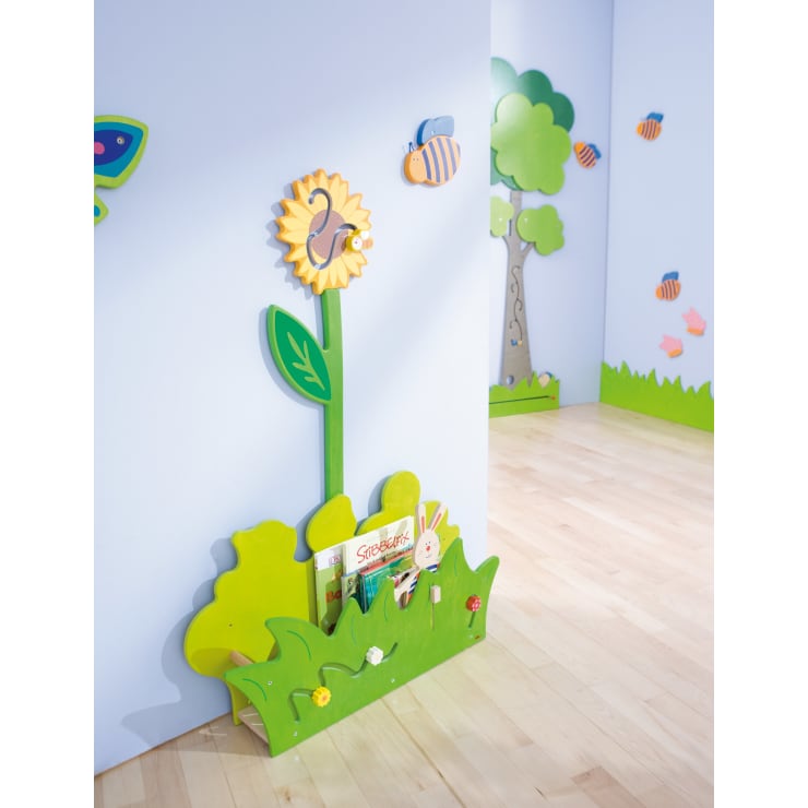 Ein farbenfrohes Kinderbuchregal in Form eines Grases und einer Sonnenblume steht vor einer hellblauen Wand und enthält ein paar Bücher. Das Zimmer hat einen Holzboden und Wanddekorationen mit Bienen, Bäumen und Blättern.