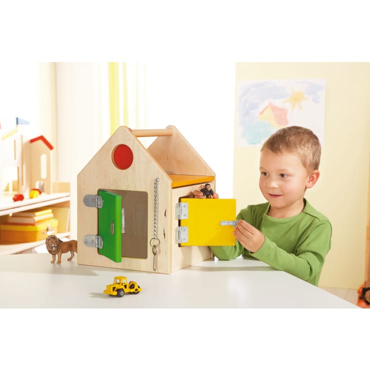 Ein kleines Kind öffnet eine gelbe Riegeltür an einem hölzernen Spielhaus mit mehreren Türen und Schlössern. Spielzeugfiguren, ein Löwe und ein gelber Lastwagen stehen auf dem Tisch. An der Wand im Hintergrund hängen Zeichnungen.