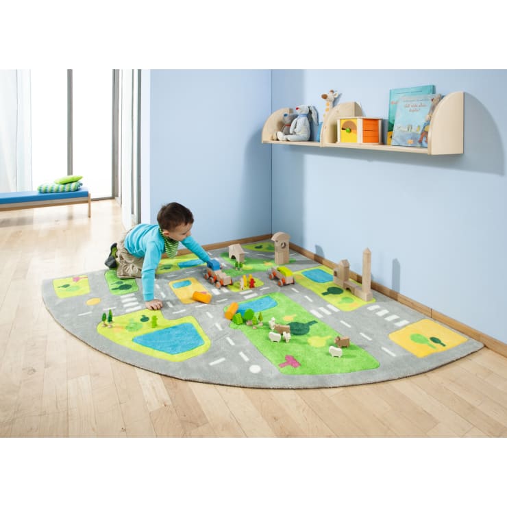 Ein kleines Kind in einem blauen Hemd spielt mit Holzklötzen und Spielzeugfahrzeugen auf einer runden, illustrierten Spielmatte in einem hellen Raum mit Holzboden und hellblauen Wänden. An der Wand dahinter befinden sich Regale mit Büchern und Spielzeug.