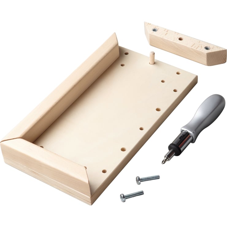 Ein rechteckiges Holzbrett mit abgewinkelten Seitenteilen, einem Holzpflock, mehreren Löchern, zwei Schrauben, einem Schraubenzieher und einem weiteren Holzstück mit zwei Löchern, angeordnet auf einer weißen Fläche.