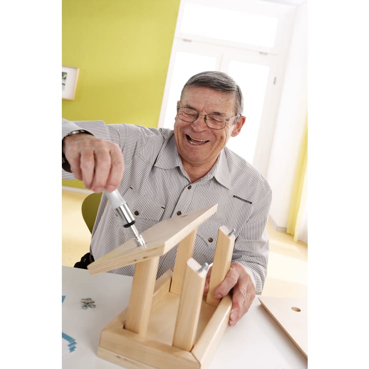 Ein älterer Mann mit Brille lächelt, während er mit einem Schraubenzieher an einem weißen Tisch in einem hellen Raum mit hellgrünen Wänden ein kleines Holzmöbelstück zusammenbaut.