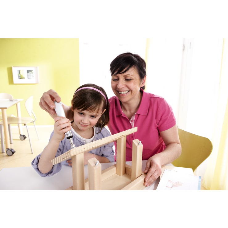 Eine erwachsene Frau und ein junges Mädchen bauen gemeinsam eine Holzkonstruktion zusammen. Das Mädchen benutzt einen Schraubenzieher, während die Frau zusieht und sie anleitet. Sie befinden sich in einem hellen, modernen Raum mit grünen und weißen Wänden.