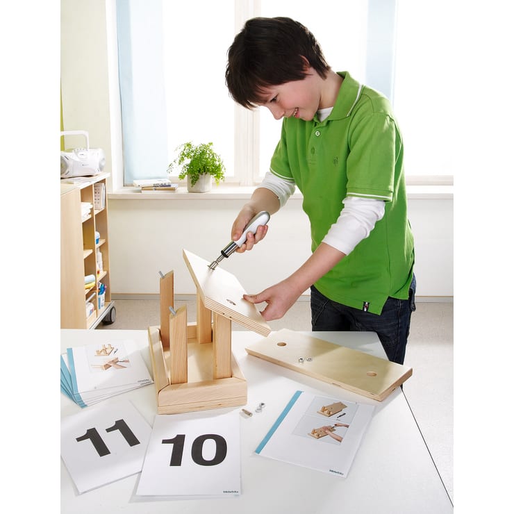 Ein Junge in einem grünen Hemd setzt mit einem Schraubenzieher eine Holzkonstruktion an einem Tisch zusammen. Auf dem Tisch liegen Anleitungshefte, nummerierte Karten und Kleinteile aus.