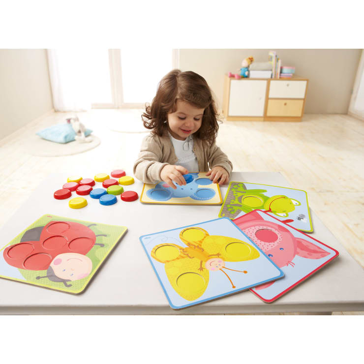 Ein kleines Kind sitzt an einem weißen Tisch und legt runde Teile in ein blaues Puzzlebrett mit Tiermotiven. Andere bunte Puzzlebretter und -teile sind auf dem Tisch verteilt. Der Raum ist hell und minimalistisch eingerichtet.