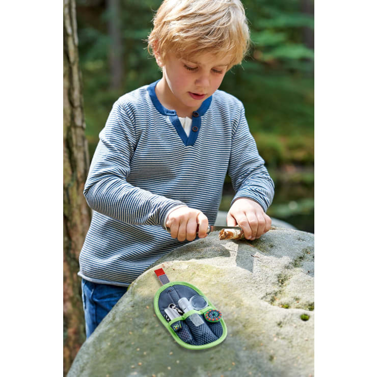 Ein kleines Kind in einem gestreiften Hemd schnitzt mit einem kleinen Messer einen Stock, während es im Freien an einem großen Felsen steht. Ein grüner Beutel mit Werkzeugen liegt auf dem Felsen. Im Hintergrund sind Bäume und Grünpflanzen zu sehen.
