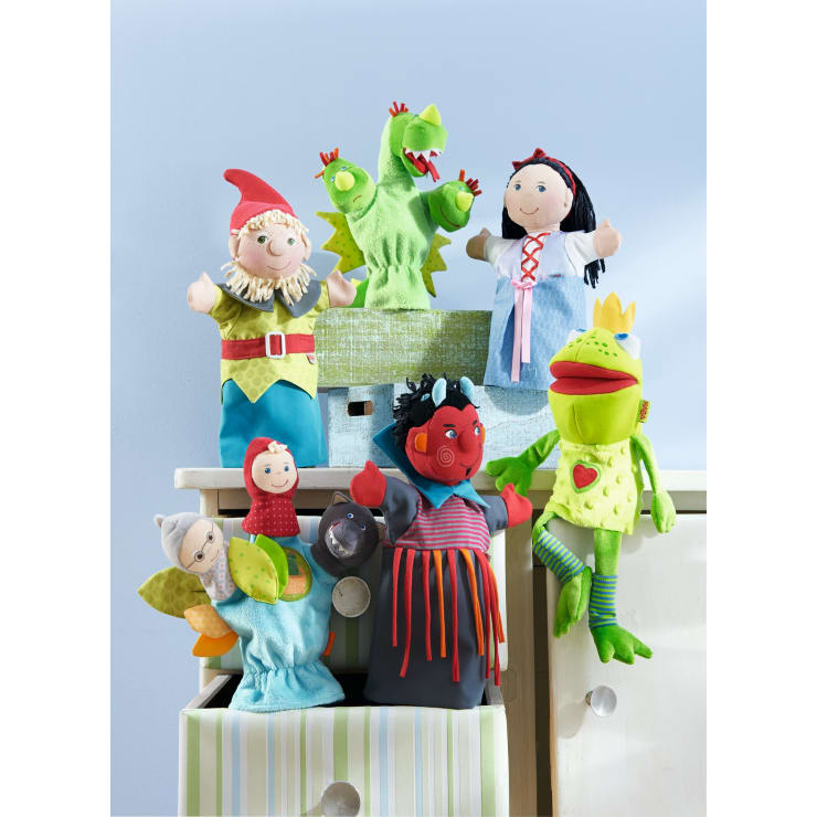 Sechs bunte Handpuppen, darunter ein Gnom, ein grüner Drache, ein Mädchen in einem blauen Kleid, ein Frosch, eine Hexe und eine Figur mit einem roten Umhang, sind auf Schubladen und Kisten vor einer hellblauen Wand angeordnet.