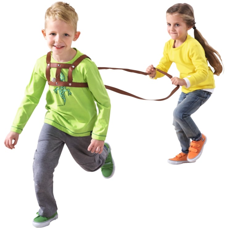 Zwei Kinder spielen. Ein Kind in grünem Hemd und grauer Hose läuft voraus, während ein anderes Kind in gelbem Hemd und Jeans sich an einem Geschirr und einer Leine festhält, die am ersten Kind befestigt sind.
