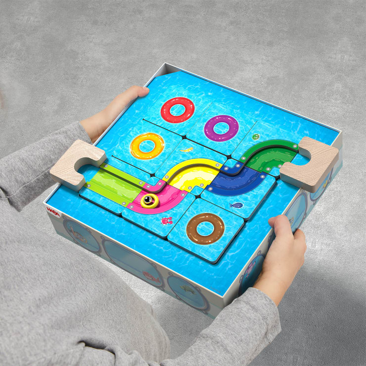 Ein Kind hält ein quadratisches, blaues Puzzlespielzeug mit bunten, geschwungenen Bahnen, Holzgriffen auf jeder Seite und kreisförmigen Öffnungen in Rot, Rosa, Gelb und Braun auf der Oberfläche. Der Hintergrund ist ein grauer, strukturierter Boden.