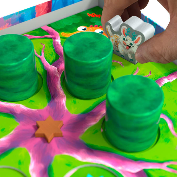 Eine Hand setzt eine weiße, tierförmige Spielfigur auf ein buntes Brett mit grünen, zylindrischen Stiften, einem rosa Verzweigungsmuster und einem orangefarbenen, sternförmigen Spielstein.