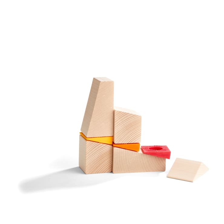 Ein kleines Arrangement aus hellen Holzklötzen und zwei farbigen Verbindungsstücken, einem orangefarbenen und einem roten, die zu einer geometrischen Struktur auf weißem Hintergrund zusammengesetzt sind. Ein Block liegt flach im Vordergrund.