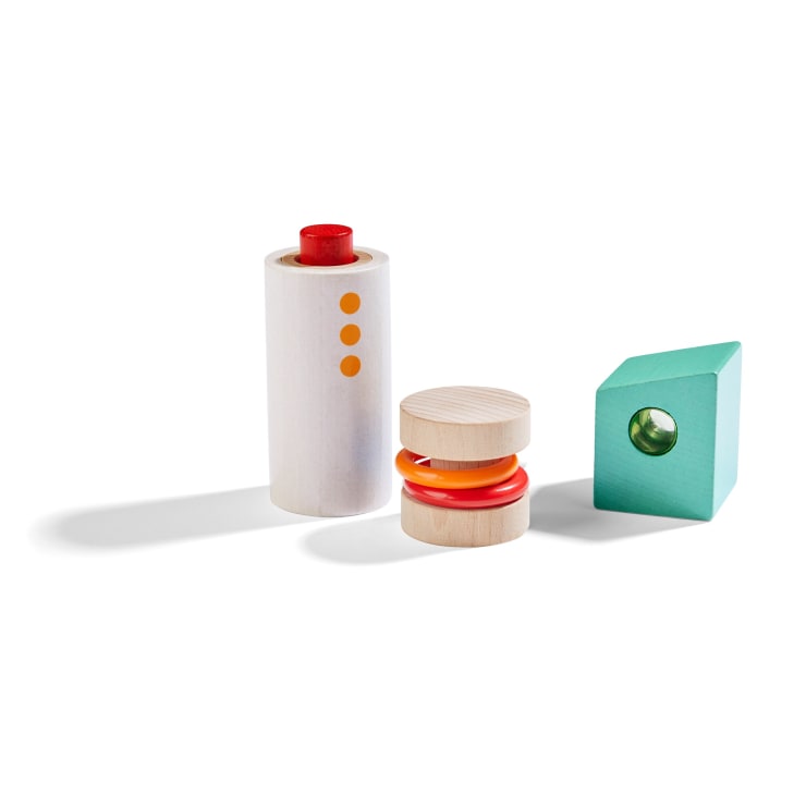 Drei Holzspielzeugformen auf weißem Hintergrund: ein hoher weißer Zylinder mit orangefarbenen Punkten und einer roten Spitze, zwei beigefarbene Scheiben mit orangefarbenen und roten Ringen dazwischen und ein grüner Block mit einem runden Loch.